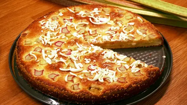 En saftig rabarberkladdkaka med smulad vit choklad och färska rabarberstjälkar i bakgrunden.