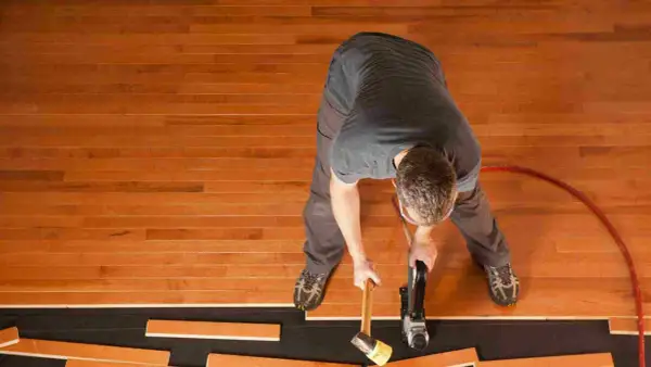 A man is installing waterproof flooring, using a nail gun and mallet to secure wooden planks.