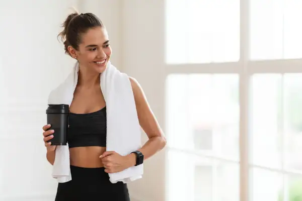 Lächelnde Frau mit Handtuch und Shaker nach dem Training. Sie fragt sich, wie viel Kreatin pro Tag für optimale Ergebnisse.