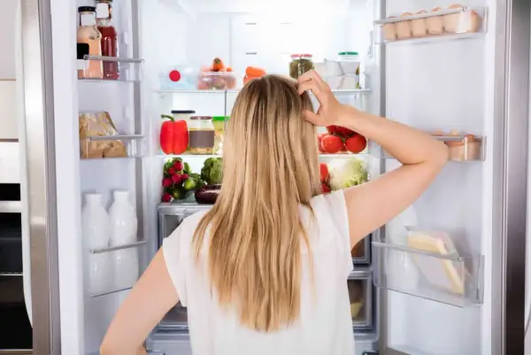 Donna confusa davanti al frigo pieno di cibo, con il desiderio di mangiare e non ingrassare, ma lo stress la blocca.