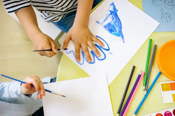 Manos de niños pintando con acuarelas y lápices de colores, aprendiendo los lomloe saberes básicos.
