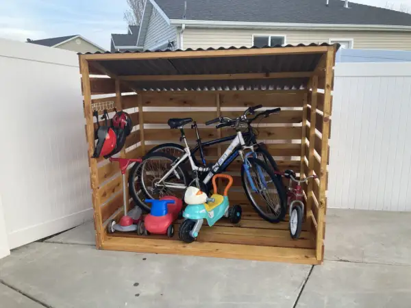 A DIY bike rack made of wood stores two adult bikes, a tricycle, and two ride-on toys. Helmets hang on a hook.
