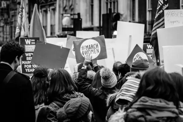 Tłum na marszu, gdzie feminizm i emancypacja idą w parze. Ludzie niosą transparenty, jeden z nich głosi "#MARCH 4WOMEN".