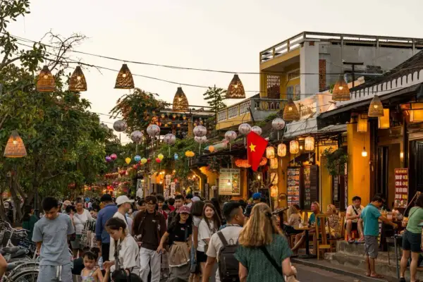 Bunte Lichter und Menschenmassen beleuchten die **Hoi An Altstadt**. Ein vietnamesisches Banner weht über einer belebten Straße voller Geschäfte und Restaurants.