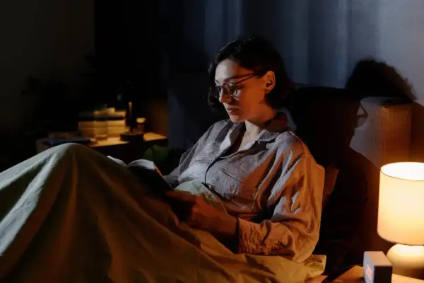 Kobieta w okularach czyta książkę w łóżku przy lampce nocnej. Czy czytanie po ciemku psuje wzrok?