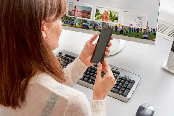 Kobieta trzyma telefon, szukając informacji, jak podłączyć telefon do monitora. Na ekranie komputera widać zdjęcia.