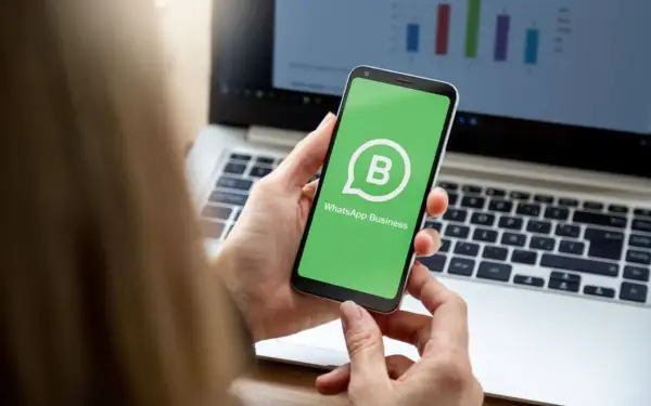 Dłoń trzyma smartfon z zielonym ekranem i logo WhatsApp Business. W tle widać laptop z wykresem.