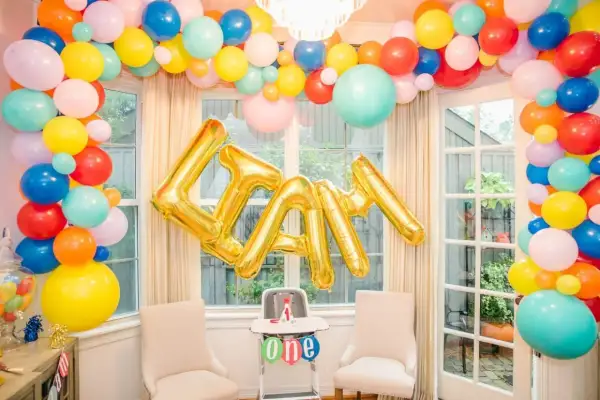Złote balony na roczek tworzą napis "LIAM" na tle kolorowej girlandy z balonów.