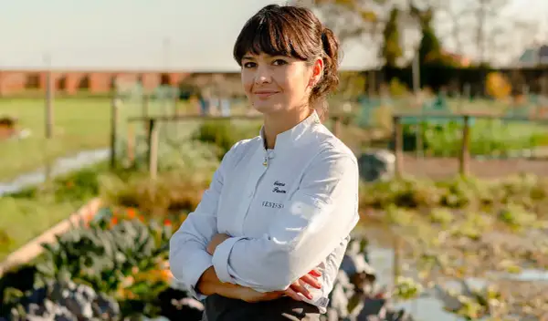 Chiara Pavan, chef con grembiule e giacca bianca, sorride con le braccia conserte in un orto.