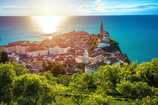 Sonnenuntergang über der malerischen Piran Altstadt, mit ihren roten Dächern und der Kirche, die auf einer Landzunge ins türkisfarbene Meer ragt.