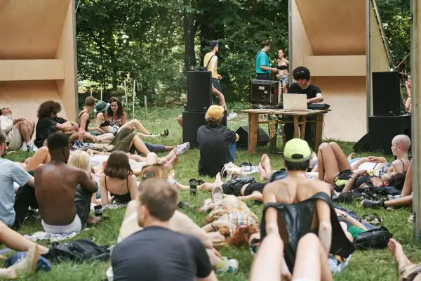Un gruppo di persone rilassate sull'erba, ascoltando musica a un butik festival. Un DJ mixa con un laptop, circondato da altoparlanti e una struttura in legno.