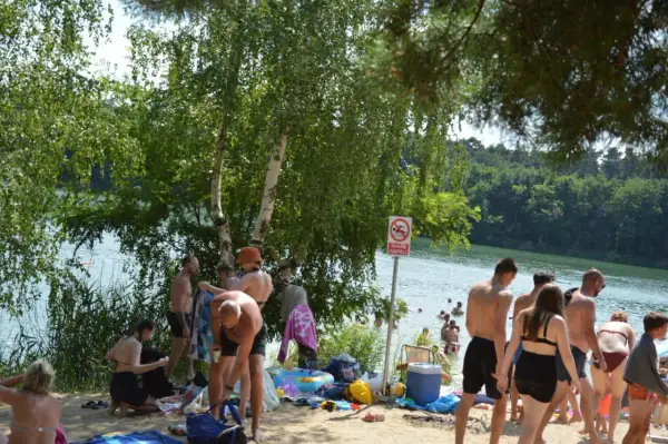 Ludzie odpoczywają nad jeziorem, niektórzy kąpią się, inni siedzą na plaży. W tle widać las. To idealne miejsce na relaks, jakby czas się zatrzymał, niczym w starożytnym rydwanie.