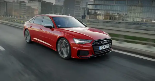 Czerwone, miękka hybryda Audi S6 sunie po drodze, za kierownicą siedzi kierowca w okularach.