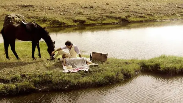 Pareja en un picnic junto a un río, con un caballo pastando. Un momento idílico para disfrutar de rutas en caballo y la naturaleza.
