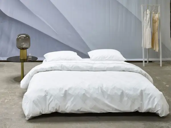 Ein Bett mit weißer Perkal-Bettwäsche, gesteppt mit feinen Linien. Daneben ein Nachttisch mit Lampe und Parfüm.