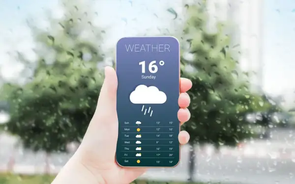 Dłoń trzyma smartfon z aplikacją pogodową pokazującą deszcz i 16 stopni. Za oknem widać krople deszczu.