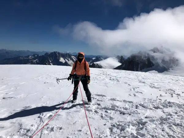 Une alpiniste, équipée pour l'ascension d'une montagne, se tient sur un sommet enneigé, le regard tourné vers l'horizon.