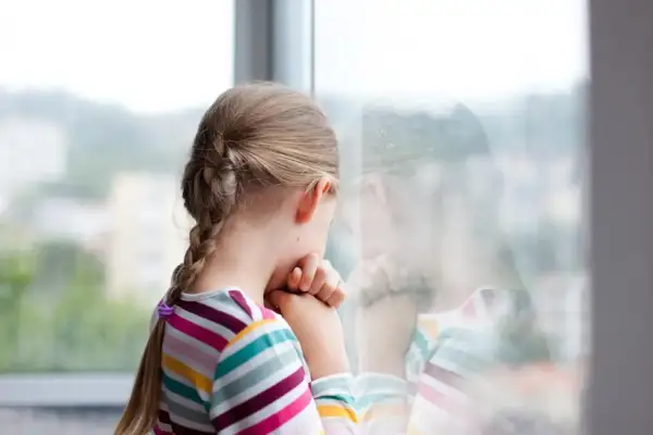Smutna dziewczynka z warkoczem patrzy przez okno. Czuje się samotna, bo dzieci nie chcą się bawić z moim dzieckiem w szkole.