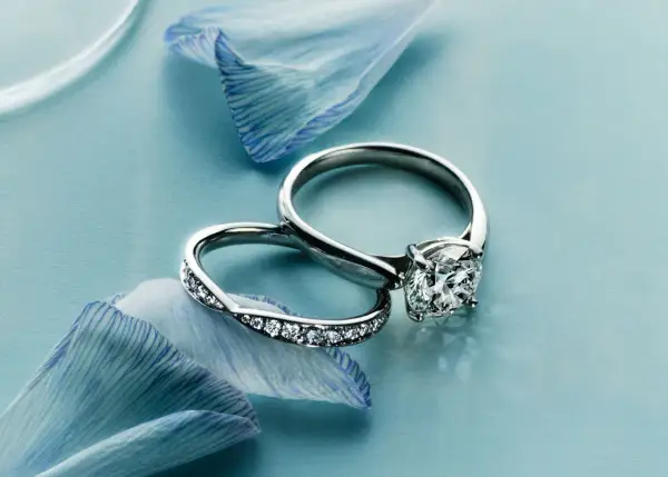 Dos anillos de compromiso, uno con un gran diamante y otro con pequeñas gemas, descansan sobre pétalos de flor azul. Un anillo casado para siempre.