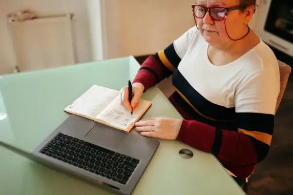 Kobieta w okularach pisze w notatniku obok laptopa.