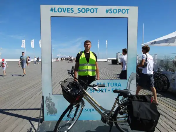 Mężczyzna w kamizelce odblaskowej pozuje przy rowerze na tle ramki z napisem #LOVES_SOPOT. Idealne miejsce na zdjęcie po zwiedzaniu tras rowerowych Sopot.