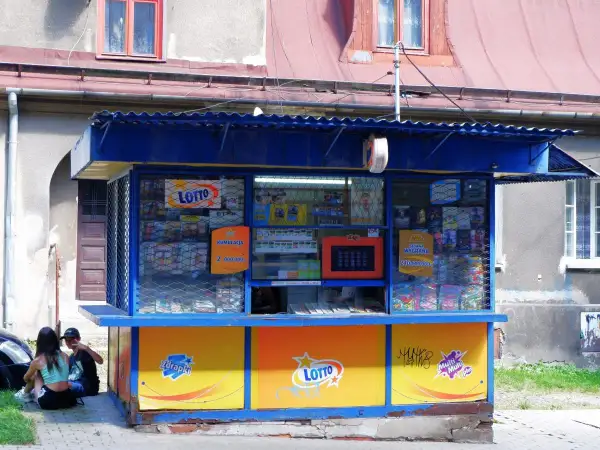Kiosk Lotto z ofertą zdrapek. Czy warto kupować zdrapki na stacjach benzynowych czy lepiej w kolekturze? Dwoje młodych ludzi siedzi obok.