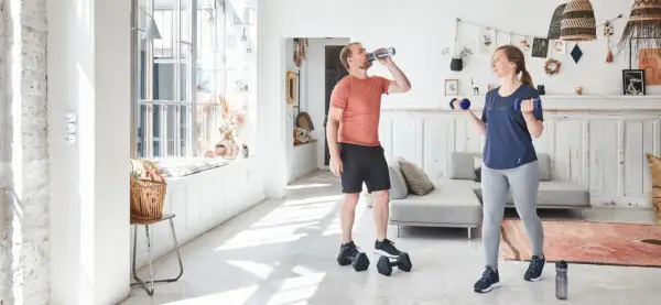 Un couple fait de l'exercice biceps maison avec des haltères bleus dans un salon lumineux.