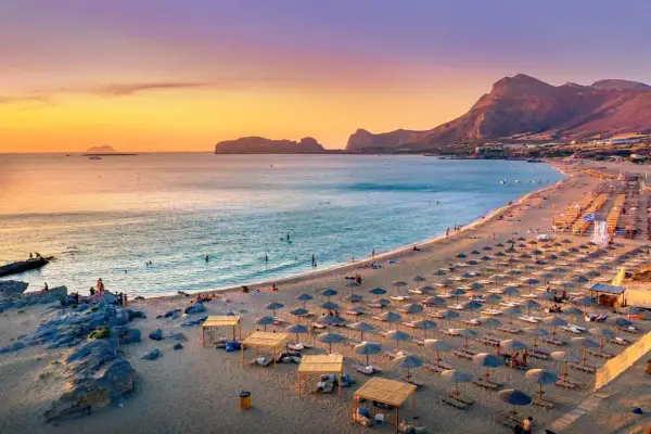 Traumhafter Badeurlaub im Mai: Sonnenschirme säumen den Sandstrand, das Meer glitzert, und die Berge im Hintergrund tauchen in warmes Abendlicht.