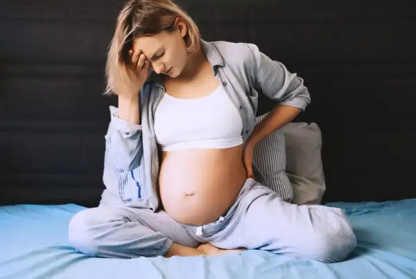 Ciężarna kobieta z bólem krocza w ciąży, trzymająca się za głowę i plecy, siedzi na łóżku.