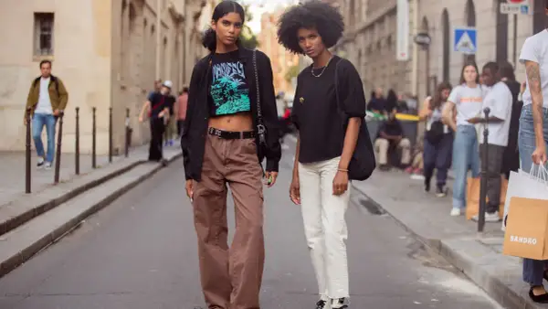 Zwei Frauen in lässiger Kleidung, eine mit brauner Cargohose und Crop-Top, die andere mit weißer Hose und schwarzem Shirt, stehen auf einer Straße.