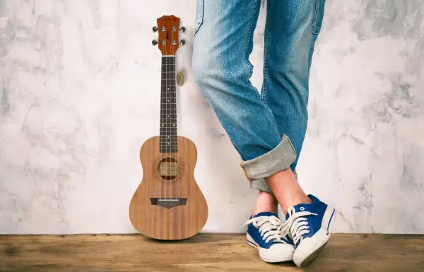 Drewniane ukulele obok osoby w jeansach i niebieskich trampkach. Idealne do grania letnich hitów na ukulele.