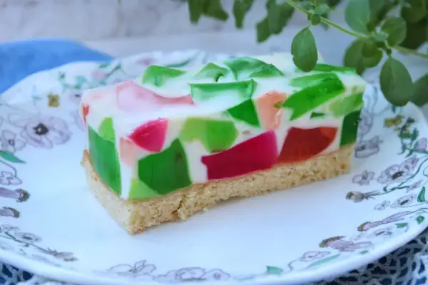 Kolorowe ciasto z galaretkami w kostkę, z zielonymi i różowymi kawałkami zatopionymi w białej masie, na spodzie biszkoptowym.
