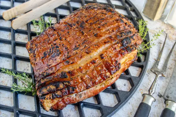 Perfekt gegrillter Schweinebauch mit knuspriger Schwarte liegt auf einem Grillrost. Kräuter und Grillbesteck deuten auf ein köstliches Mahl hin.