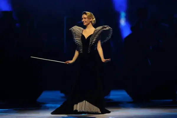 Matilda De Angelis in einem dramatischen schwarzen Kleid mit auffälligen Schulterdetails und einem Dirigentenstab.