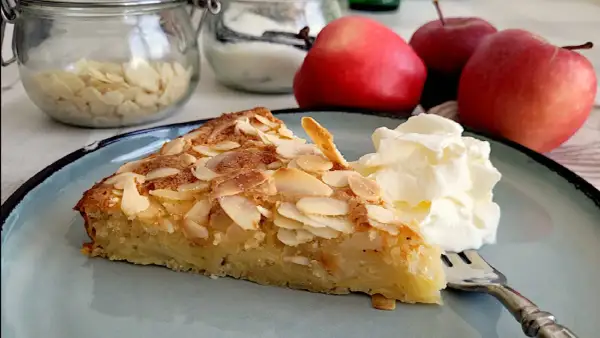 Ein Stück Apfelkuchen mit mandelkruste, garniert mit Sahne, auf einem blauen Teller. Im Hintergrund Äpfel und Mandelblättchen.
