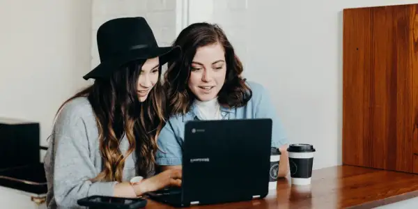 Två unga kvinnor diskuterar sin skuldkvot och bolån framför en laptop, med kaffekoppar på bordet.