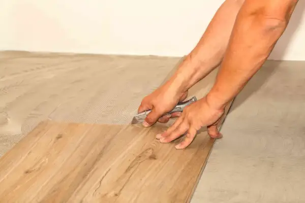 Hands using a utility knife to score and cut a vinyl plank flooring piece, demonstrating how to cut vinyl plank flooring for a perfect fit.