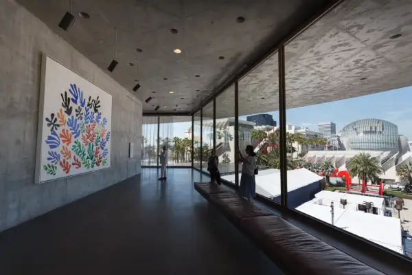 Inside a modern art museum, a colorful Matisse cutout hangs on a concrete wall. Visitors admire the art, enjoying the view of the city skyline.