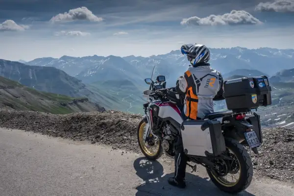 Motocyklista w kasku i kurtce podziwia górską panoramę Alp z krętą drogą w dole.