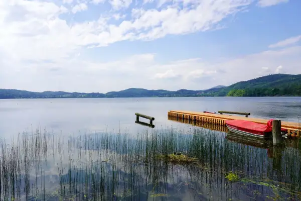 Ein Steg mit einem roten Boot am Laacher See. Schilf ragt aus dem ruhigen Wasser, das die bewaldeten Hügel am Horizont spiegelt.