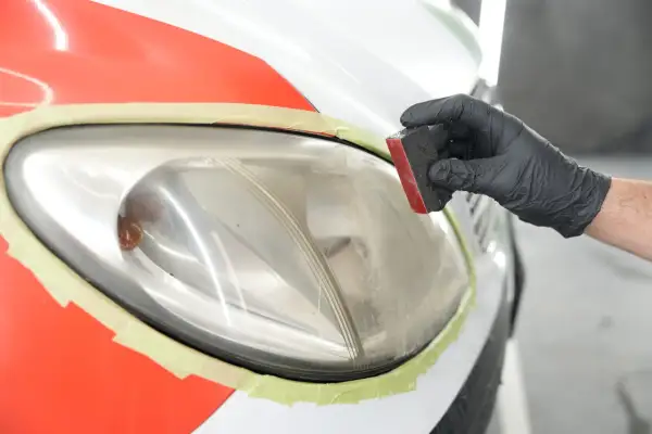 Mano con guanto nero lucida un faro auto opaco, pronto per verniciare. Nastro protettivo arancione e verde.