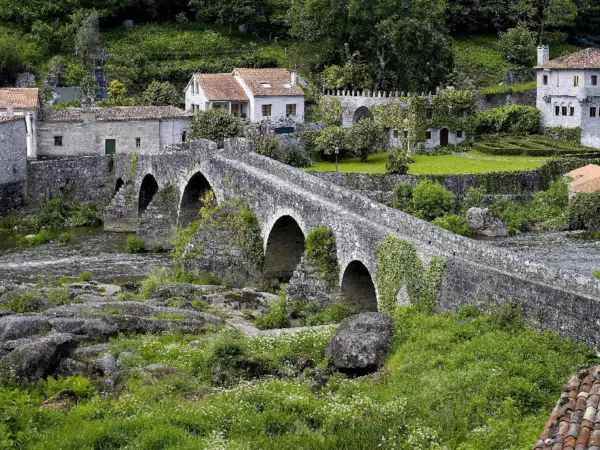 Un antiguo ponte carreira de piedra cruza un río, rodeado de casas rústicas y vegetación exuberante.