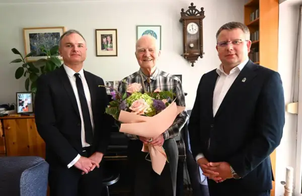 Trzech mężczyzn, jeden z bukietem kwiatów, w eleganckich strojach.