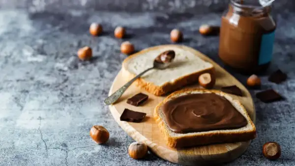 Tranche de pain généreusement tartinée de pâte à tartiner maison 3 ingrédients. Des noisettes et des carrés de chocolat parsèment la table.
