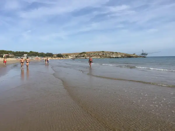 Persone passeggiano sulla battigia di Cala Molinella, con un trabucco sullo sfondo.