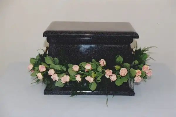 Czarna, kamienna urna ozdobiona girlandą z różowych różyczek i zieleni. Piękne kwiaty na trumnę, symbolizujące pamięć i miłość.