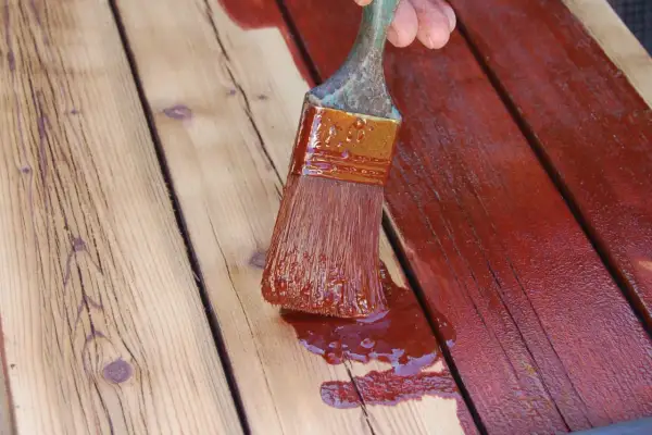 Application d'une teinture rouge sur des planches de bois. Un pinceau chargé de produit montre comment peindre du bois pour le protéger.