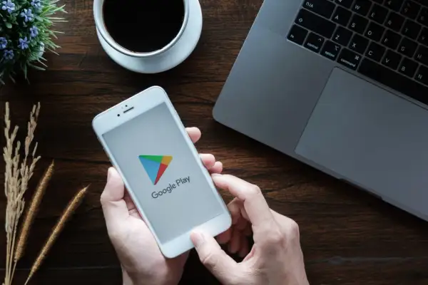 Ręce trzymają smartfon z logo Google Play, gotowe by dowiedzieć się, jak zaktualizować usługi Google Play. Obok laptop i kawa.