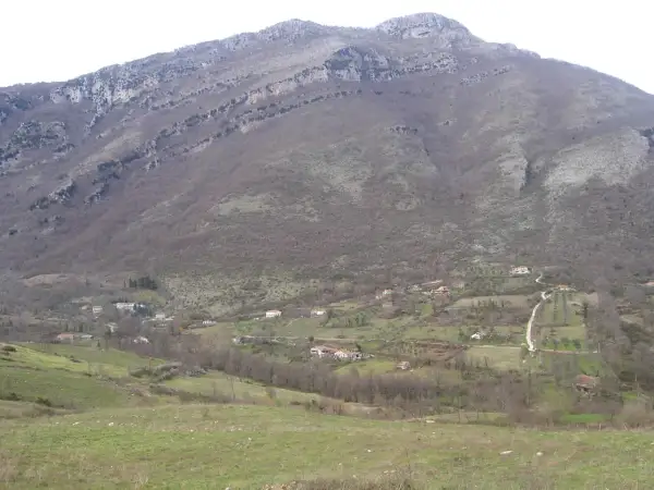 Un paesaggio collinare dove il Cilento mostra case sparse tra alberi spogli e prati verdi, sotto un imponente monte roccioso.