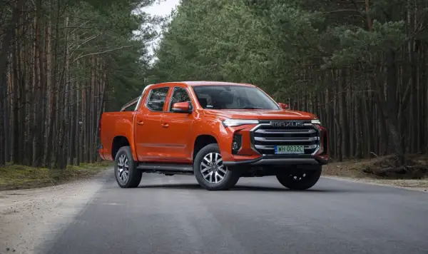 Pomarańczowy Maxus T90 na leśnej drodze. Ten potężny pickup z charakterystycznym grillem i nowoczesnymi światłami świetnie prezentuje się w otoczeniu drzew.